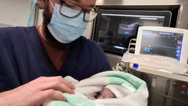 Tyrone in hospital scrubs, holding his newborn daughter swaddled in a striped blanket.
